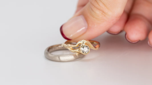 To vielsesringe ligger centralt på hvid baggrund, en herre-vielsesring i hvidguld ligger nederst og en hånd med fransk rød neglelak placerer en guld-vielsesring med stor diamant ovenpå den første.