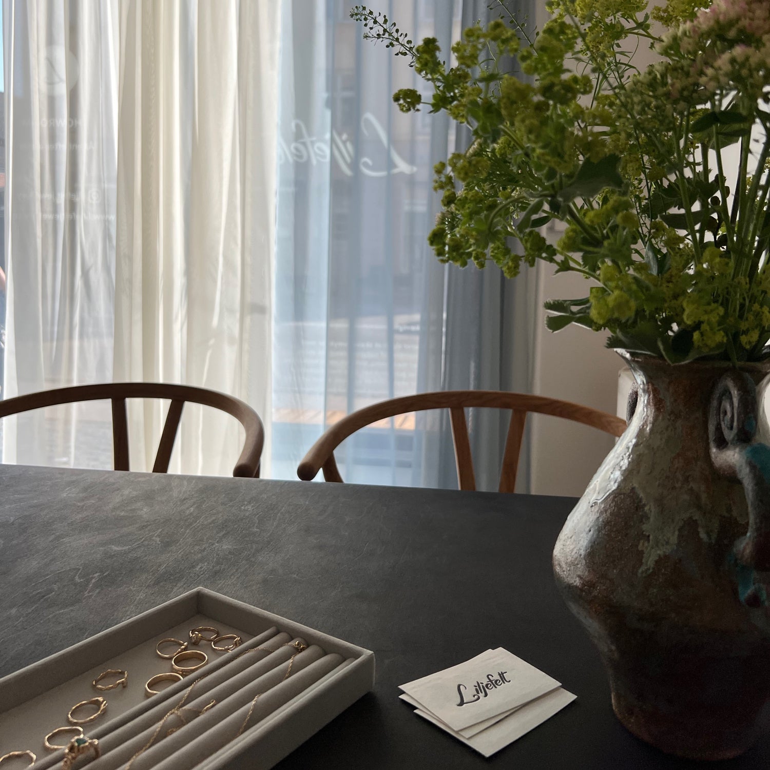 et billede fra liljefelt showroom hvor det bløde lys udefra kommer igennem de semitransperente gardiner. på bordet ligger der en række guldringe og halskæder samt liljefelt visitkort foruden en flot vase med friske blomster og to egetræs-designstole
