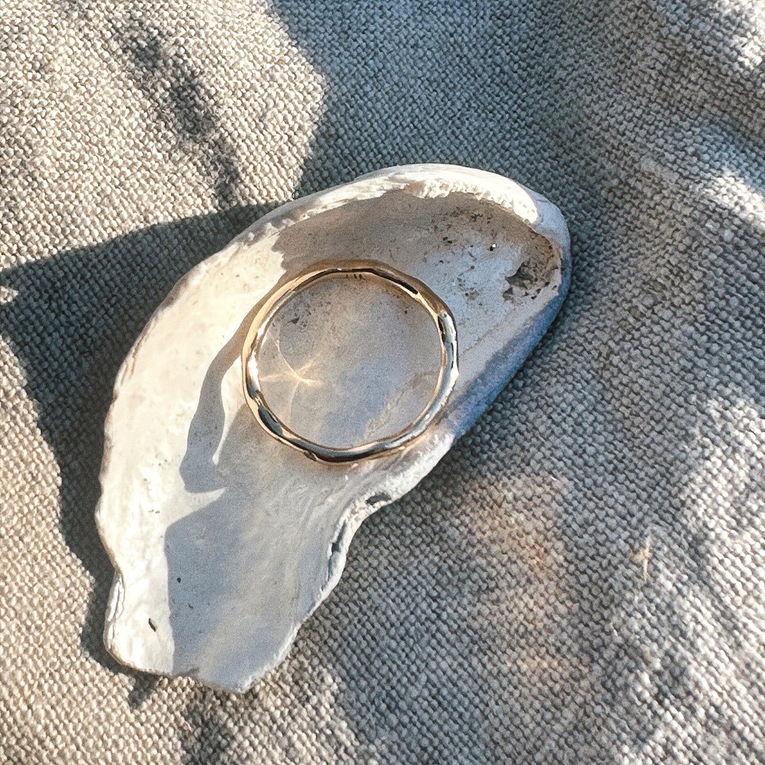 en vielsesring i guld ligger på en muslinge skal ovenpå beige stof med direkte sollys der rammer ringen og giver flotte spejlinger