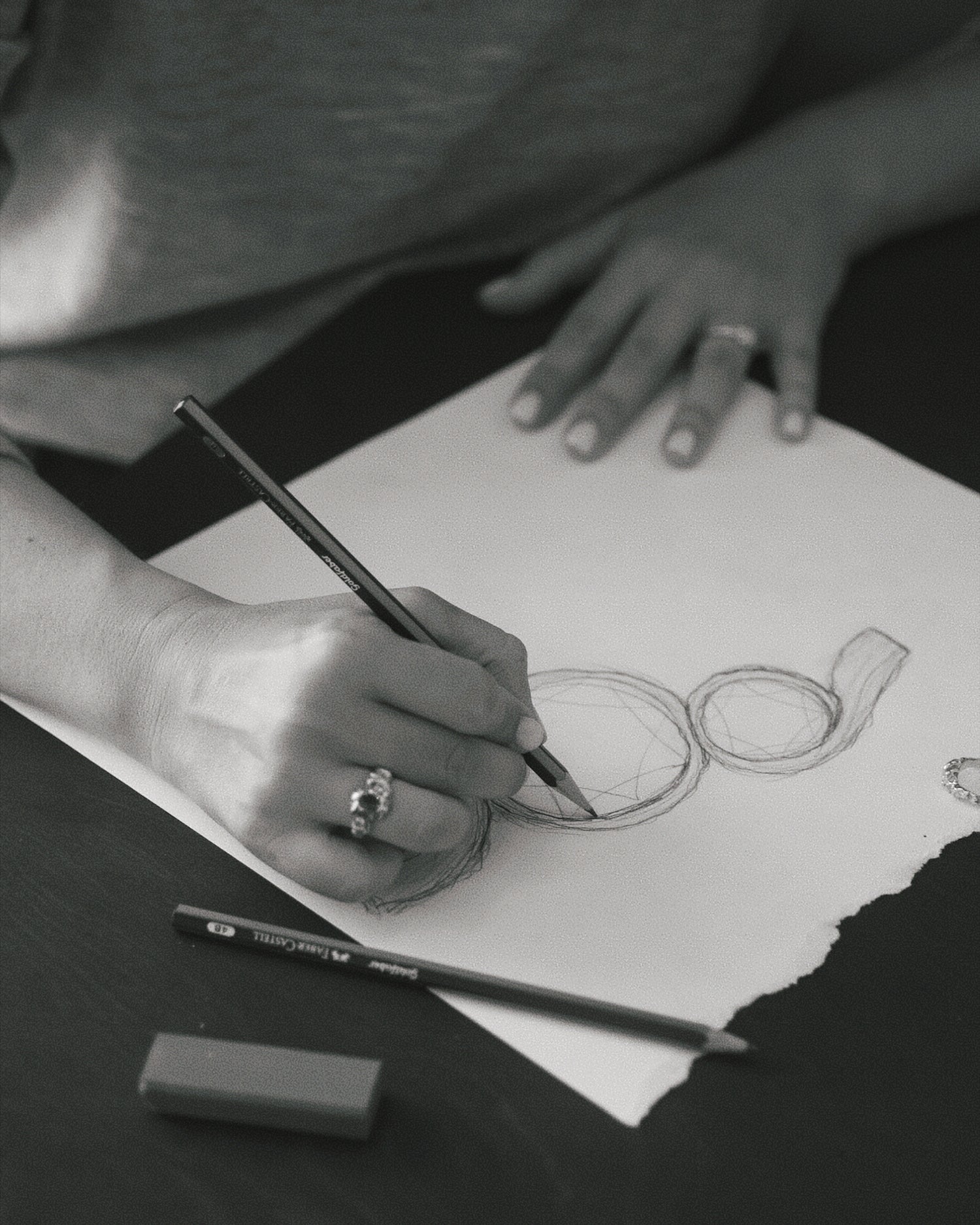 design processen af smykkedesigner michell liljefelt der tegner et forstørret ring og diamanter med en blyant. hun har en stor guldring på højre hånd med en rubin.