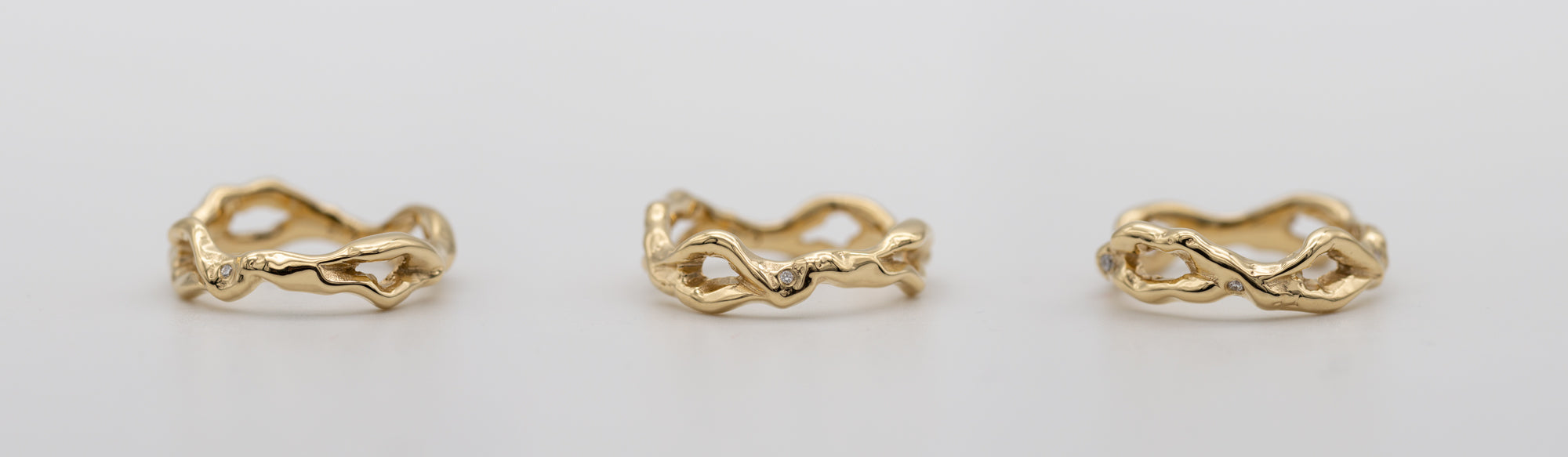 En guldring med diamanter taget fra tre vinkler ligger på grå baggrund. Ringen er i 14 karat genanvendt guld og formgivet organisk med huller i formgivningen hele vejen rundt.