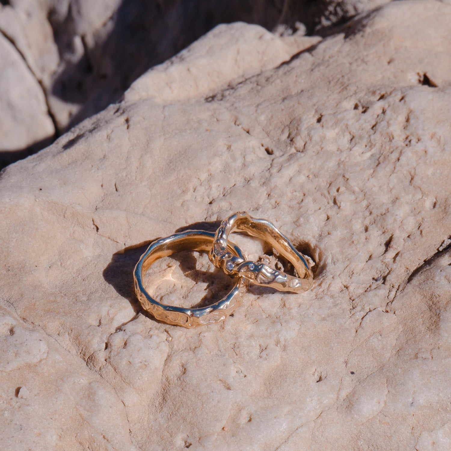 to vielsesringe ligger op ad hinanden på en klippe med direkte sollys på. Ringene er begge i genanvendt guld med diamanter der glimter