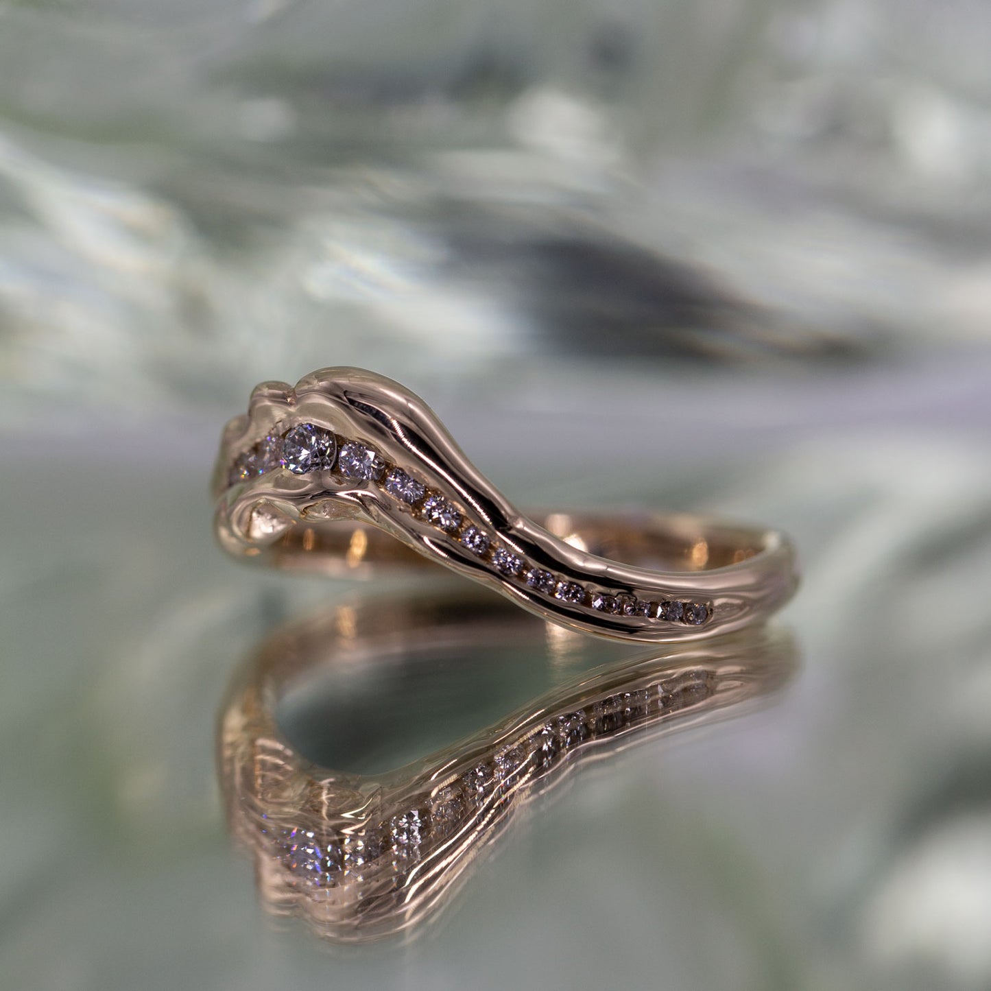 Ring i guld set i perspektiv på spejlende grøn baggrund. Ringen i guld er formgivet organisk med mange diamanter i et forløb der bugter sig som en bakketop. I midten er diamanterne størst og bliver mindre ud i mod siderne.