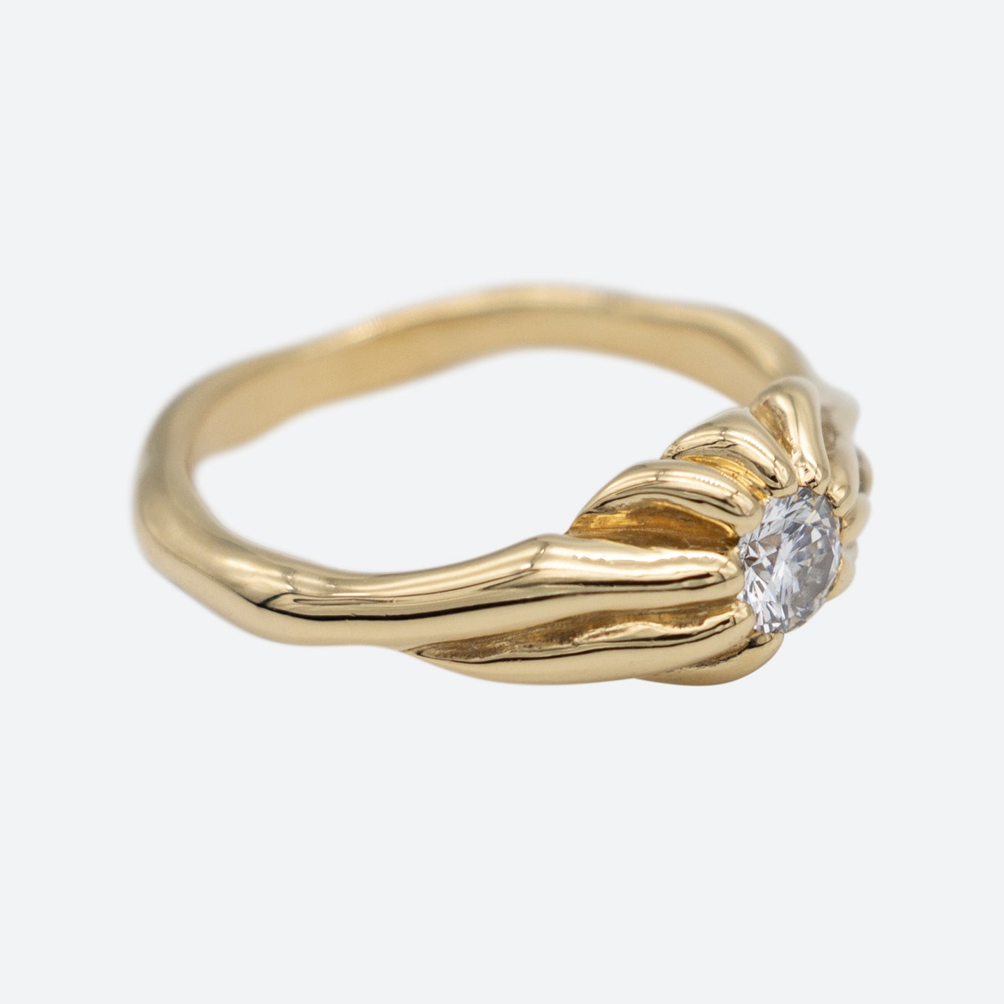 Ring i perspektiv lavet af guld med stor diamant på hvid baggrund. Guldet er formgivet som rødder der mødes i midten hvor en stor brilliantslebet diamant er placeret i midten. Design af Michell Liljefelt