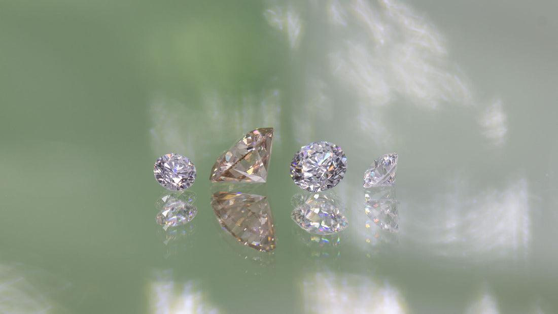 Fire diamanter i forskellige farver og størrelser ligger på række på en spejlende overflade med et grønt skær. 