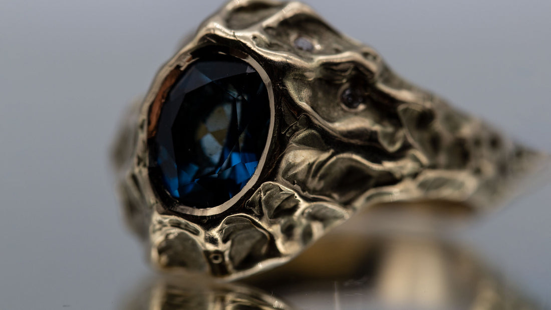 En stor guldring med en blå topas er fotograferet tæt på. Ligger på en spejlende overflade. 