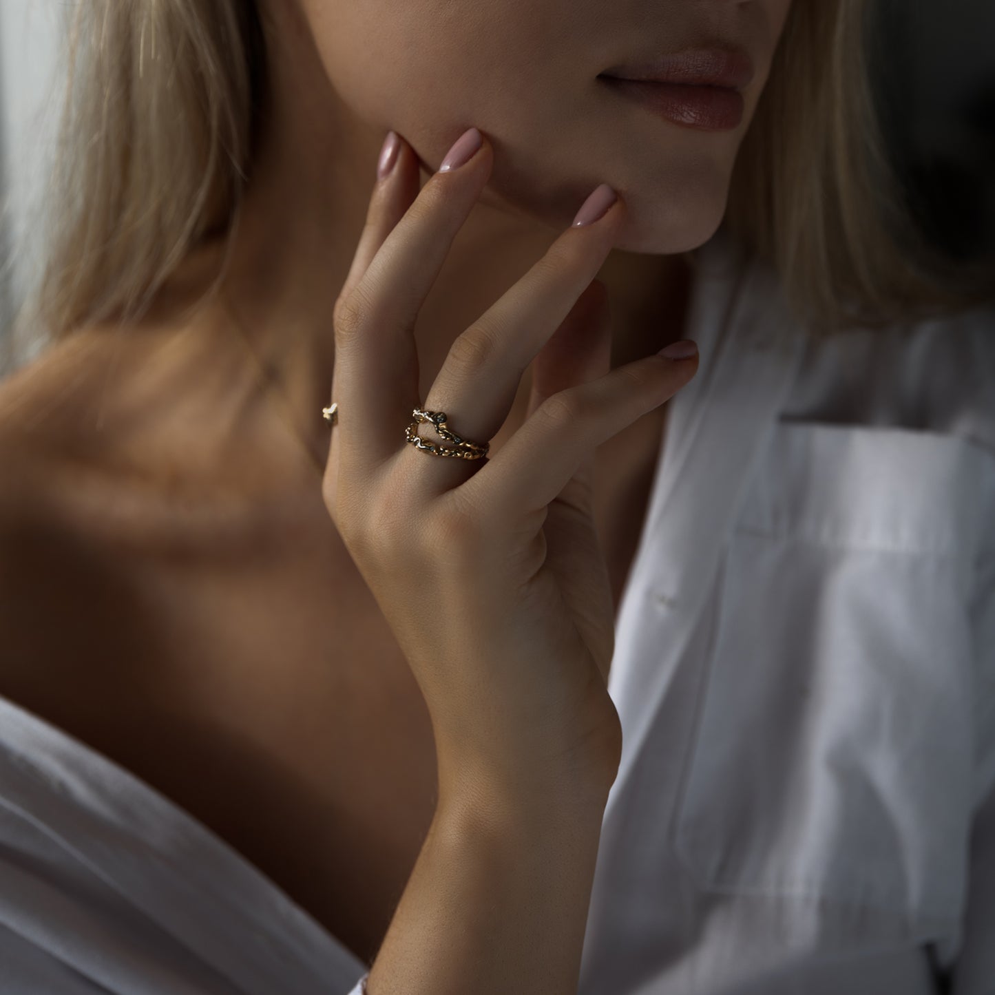 Model kvinde udsnit af hånd og kæbe, hånd holder kæben og på hånden er der to organisk designet guld ringe med diamanter der glimter. Kvinden har en hvid skjorte på der er åben foran.  