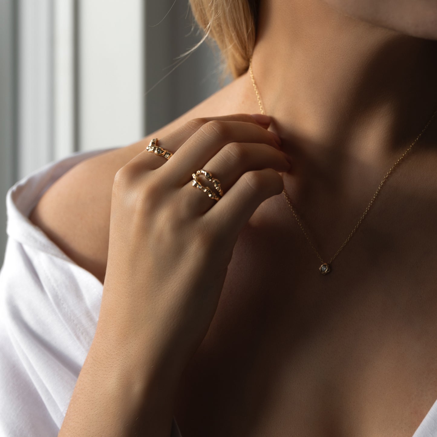 Model udsnit af torso, hånd holder guld halskæde og på hånden er der to organisk designet guld ringe med diamanter der glimter. Kvinden har en hvid skjorte på der er åben foran.  