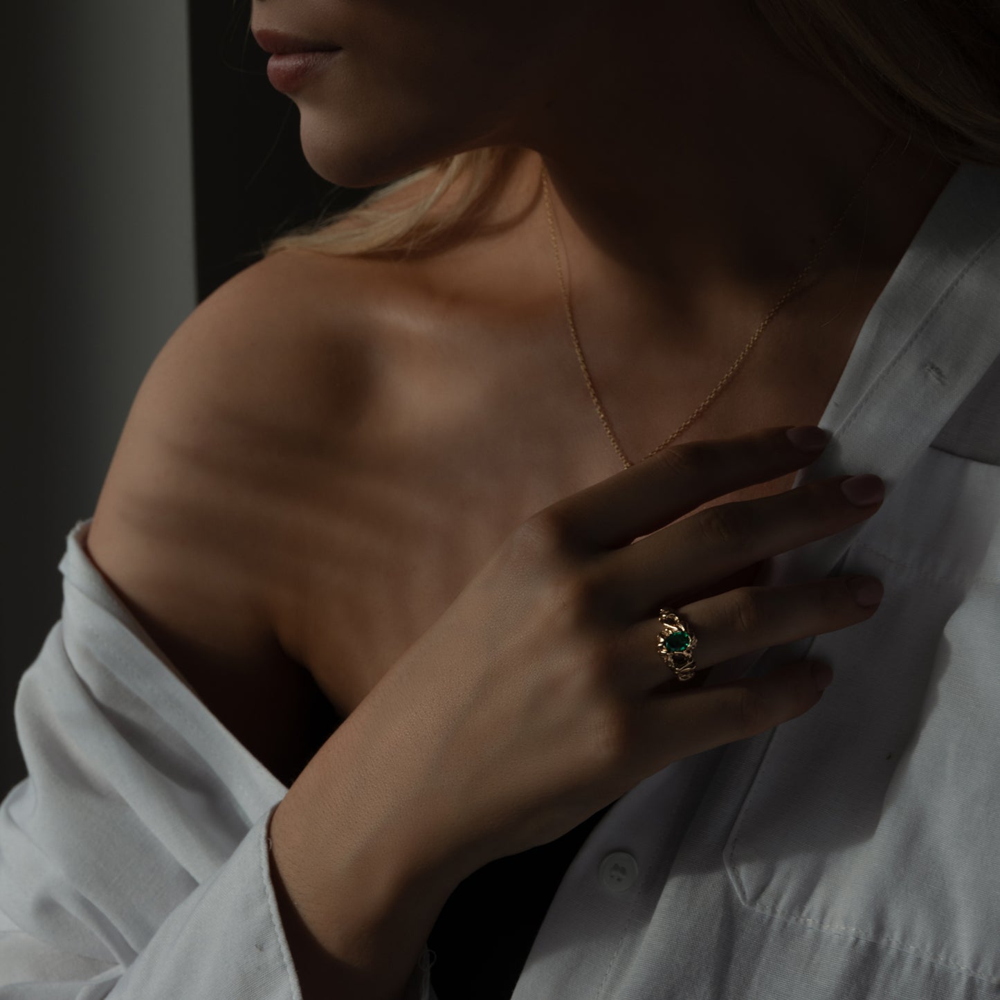 Modelbillede af torso med hånd på brystkassen, og langt lyst hår. På hånden sidder en stor organisk formgivet ring med en grøn smaragd der glimter. Til venstre for er en guld halskæde med lav dybdeskarphed.