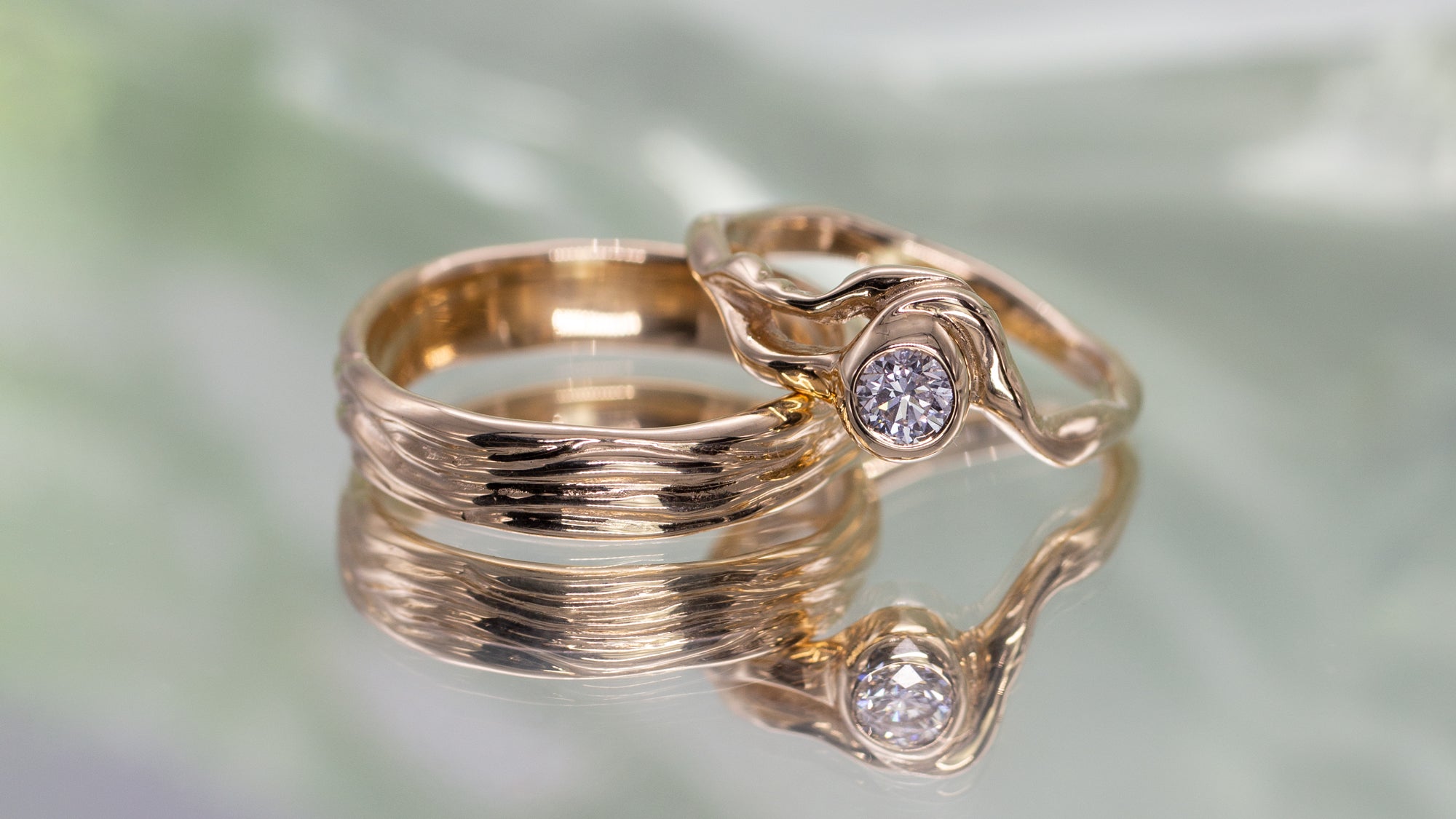 To vielsesringe i genanvendt guld. Begge er organisk formgivet. Ringen til venstre har horisontale linjer der minder om drivtømmer. Ringen til højre er med en stor fairtrade diamant i midten. 