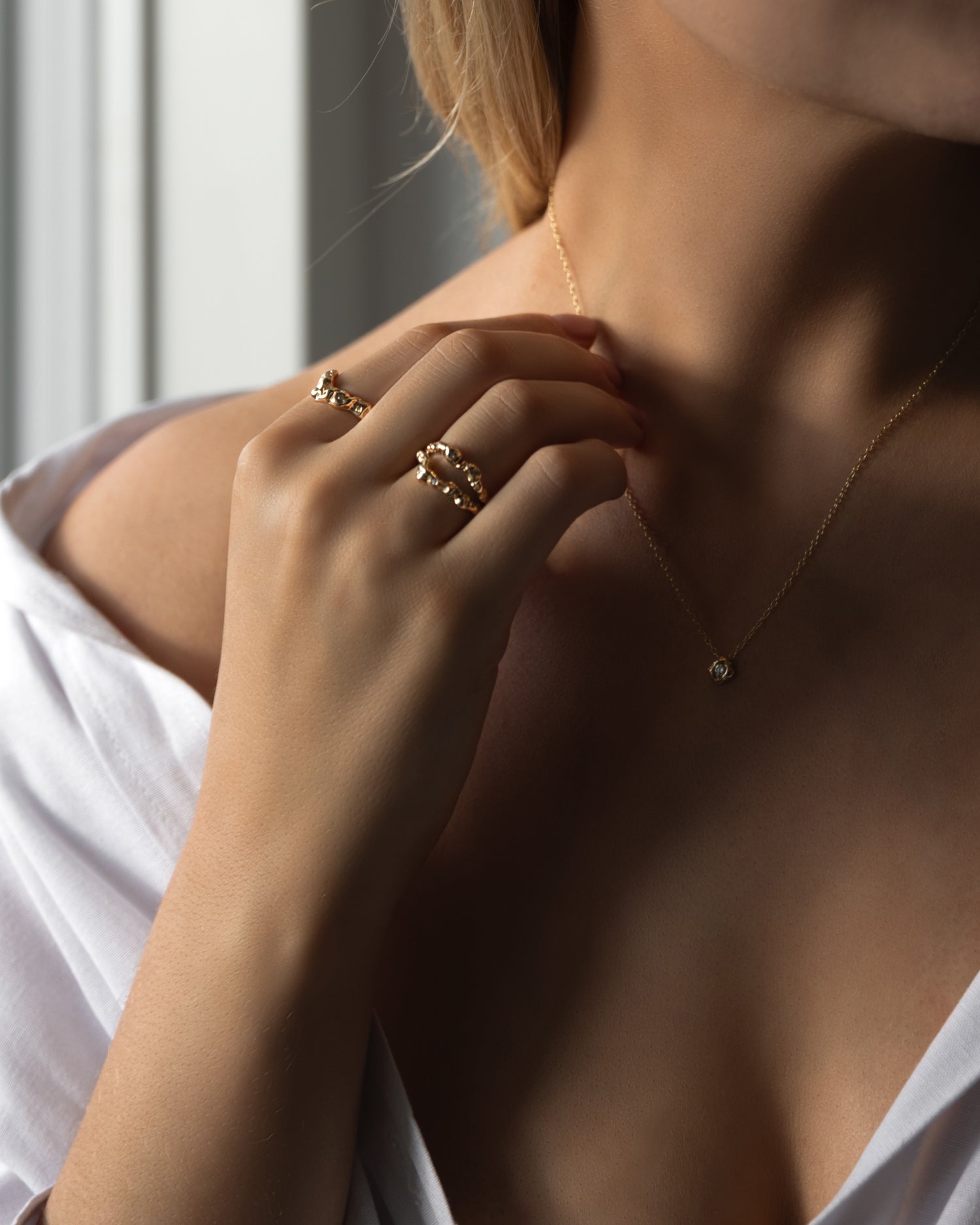 Flot kvinde udsnit af torso, hånd holder guld halskæde og på hånden er der to organisk designet guld ringe med diamanter der glimter. Kvinden har en hvid skjorte på der er åben foran.  