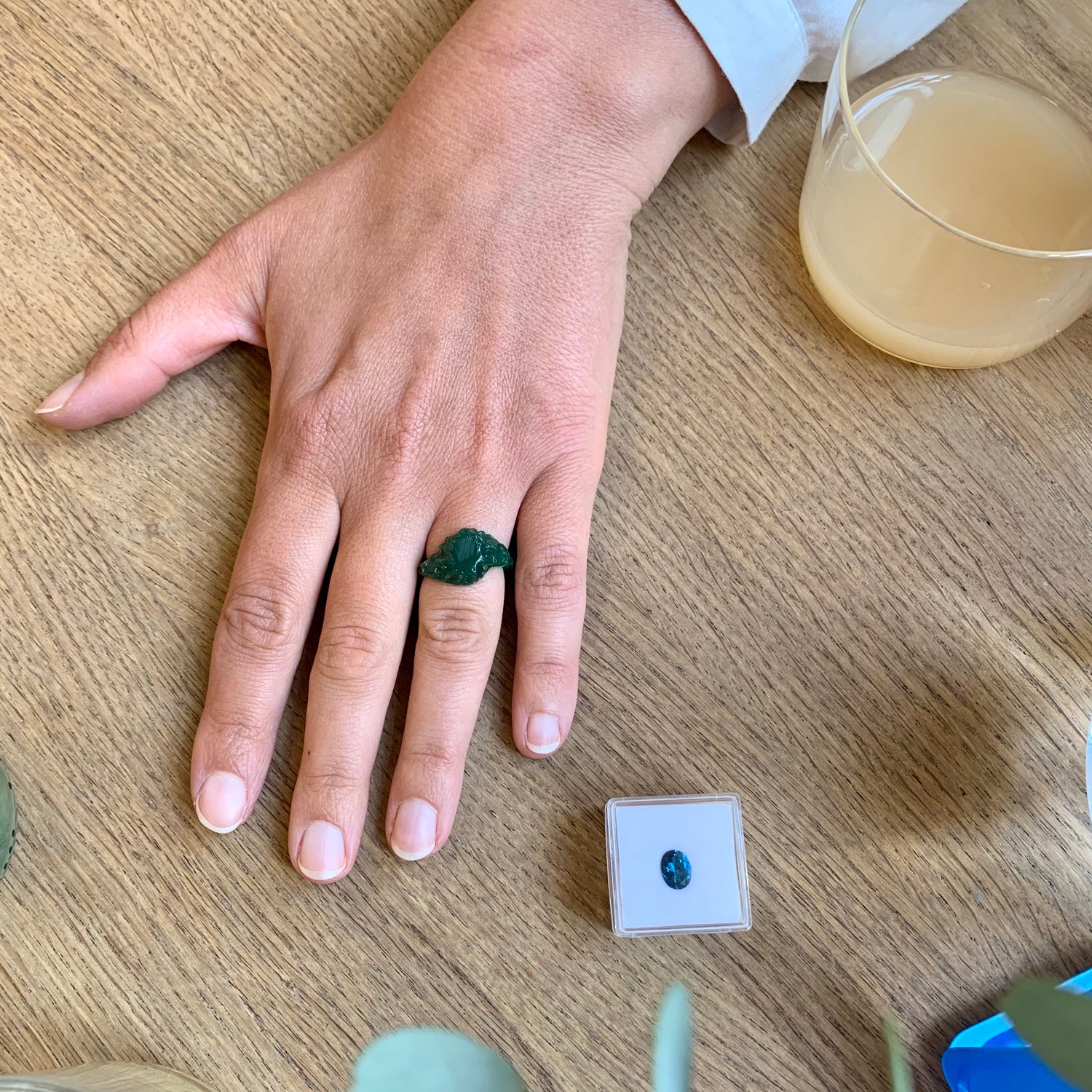 Et glas kombucha og en hånd med en ring i voksmodel og en blå topas ligger ved siden af på et egetræsbord. Design af Michell Liljefelt