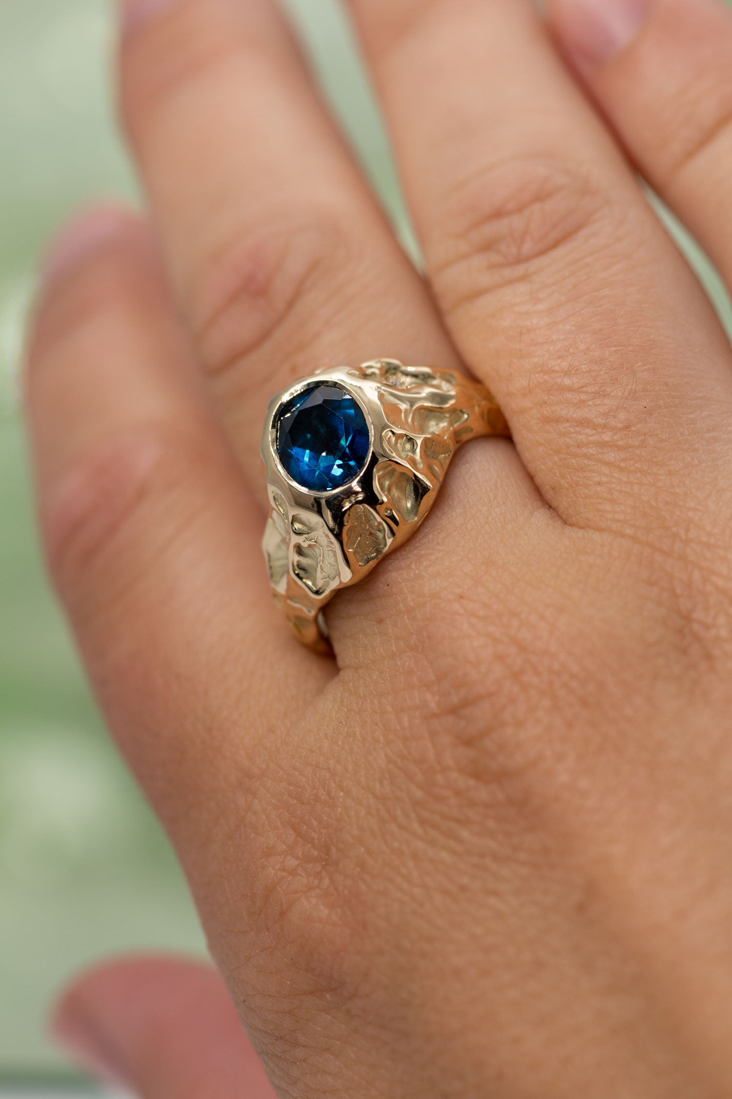 Udsnit af hånd med ring. En stor guldring sidder på pegefingeren. Ringen er med en blå topas og enkelte diamanter langs siden. Den er formgivet i et organisk udtryk. Design af Michell Liljefelt