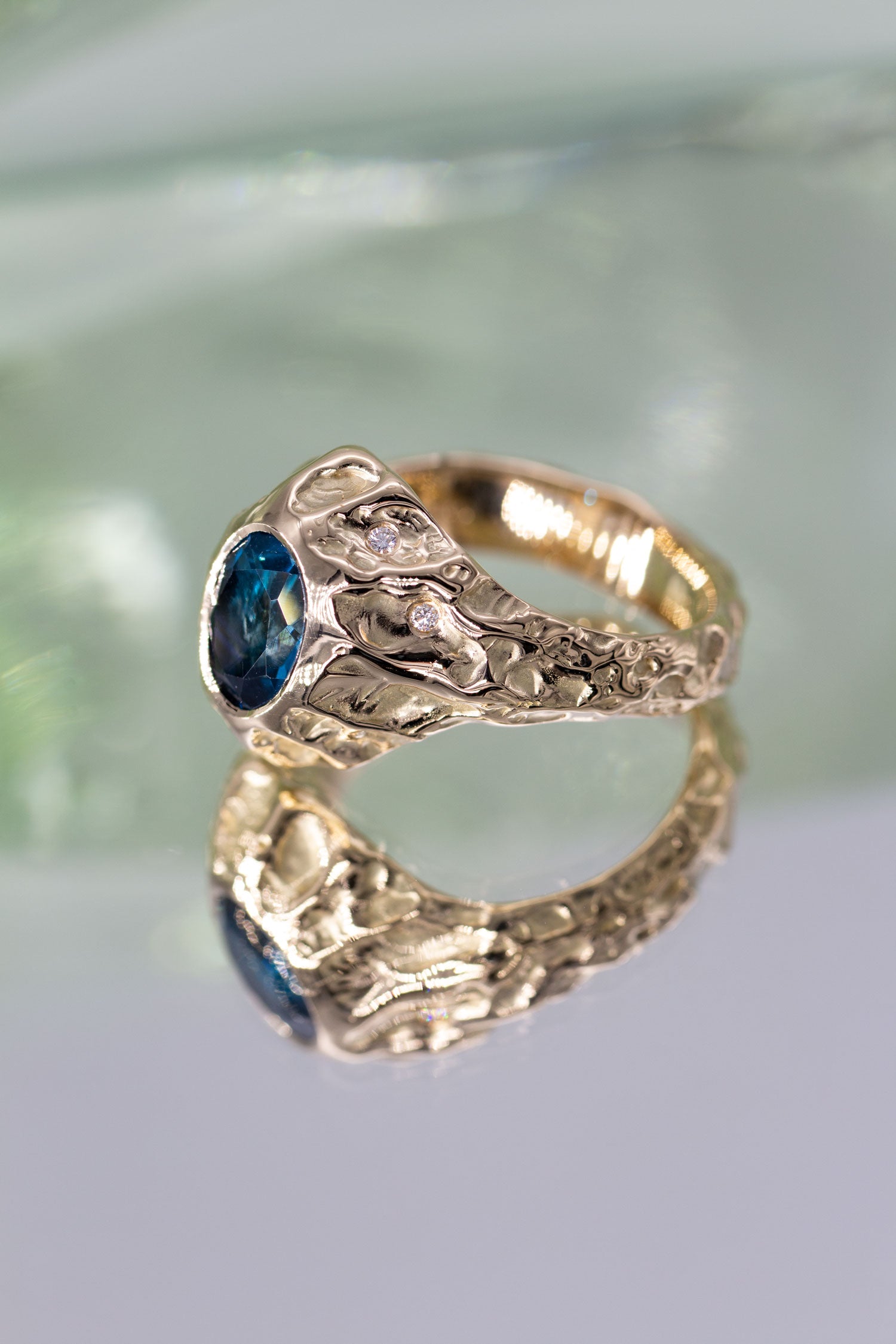 En stor guldring ligger med siden til på en spejlende, grønlig overflade. Ringen er med en blå topas og enkelte diamanter langs siden. Den er formgivet i et organisk udtryk. Design af Michell Liljefelt