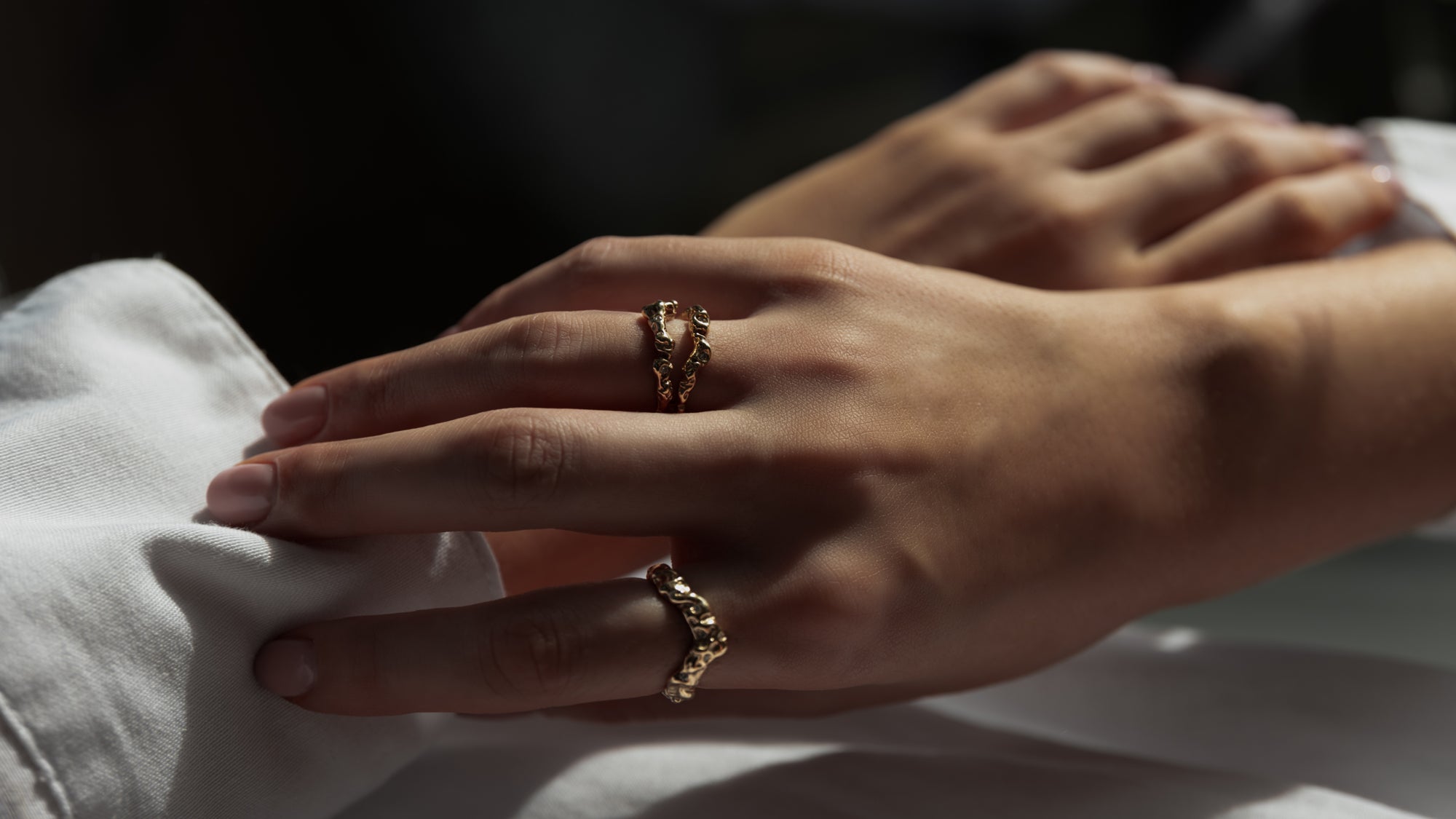 to hænder, med den højre over den venstre, hvorpå der er to organisk designet ringe på. Begge ringe er i guld med diamanter der glimter. Sort baggrund og hvid skjorteærmer 