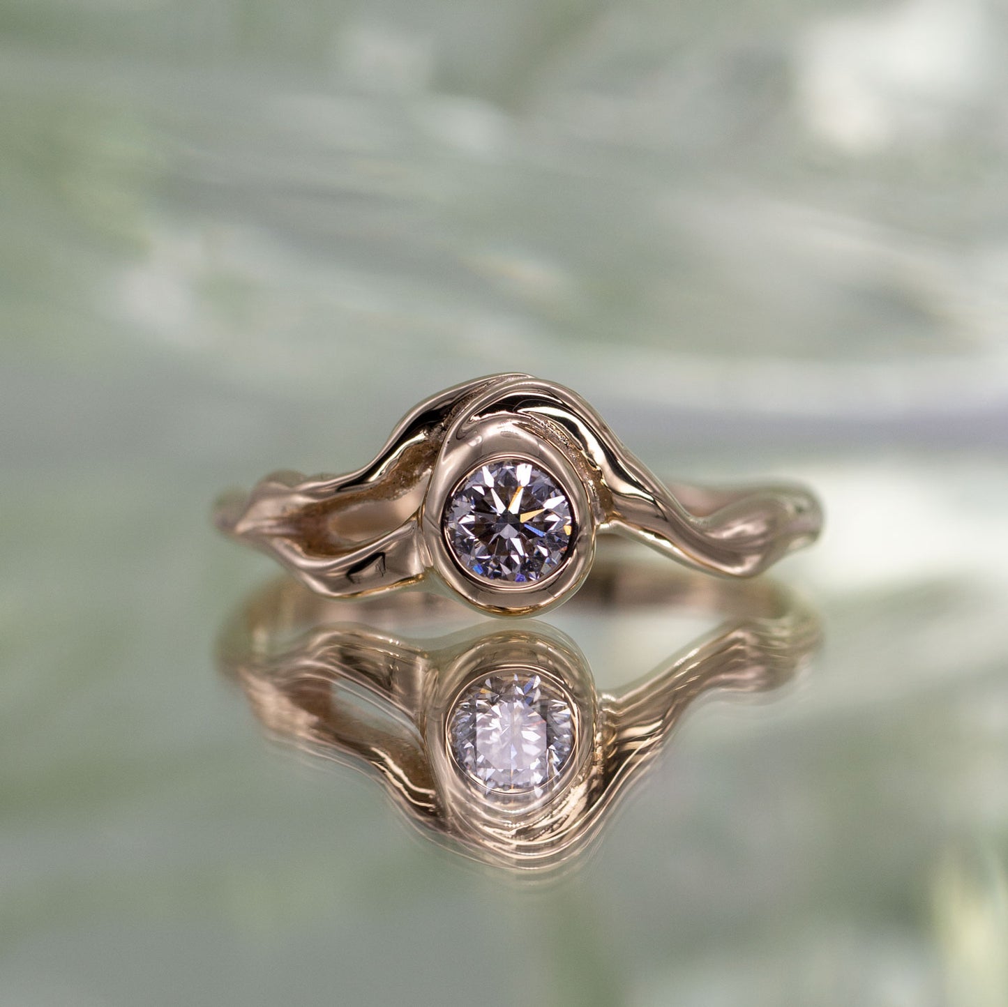 Organisk formgivet ring i guld med stor diamant placeret i centrum af ringen på spejlende lysegrøn baggrund. Ud fra diamanten forgrener formgivningen sig rundt i ring skiven. Set forfra. 