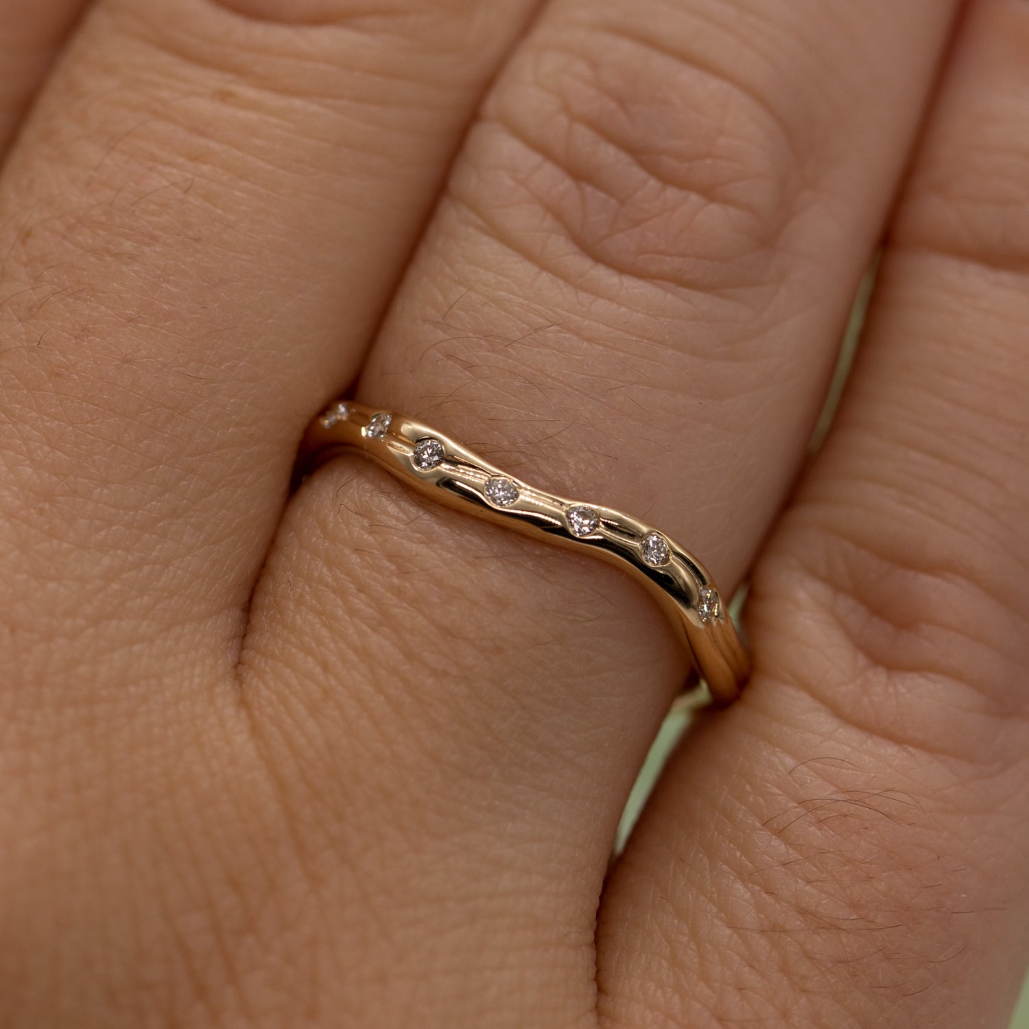 Udsnit af hånd set tæt på der viser en ring i guld med syv diamanter på lysegrøn baggrund. Ringen er formgivet organisk og bølger elegant.