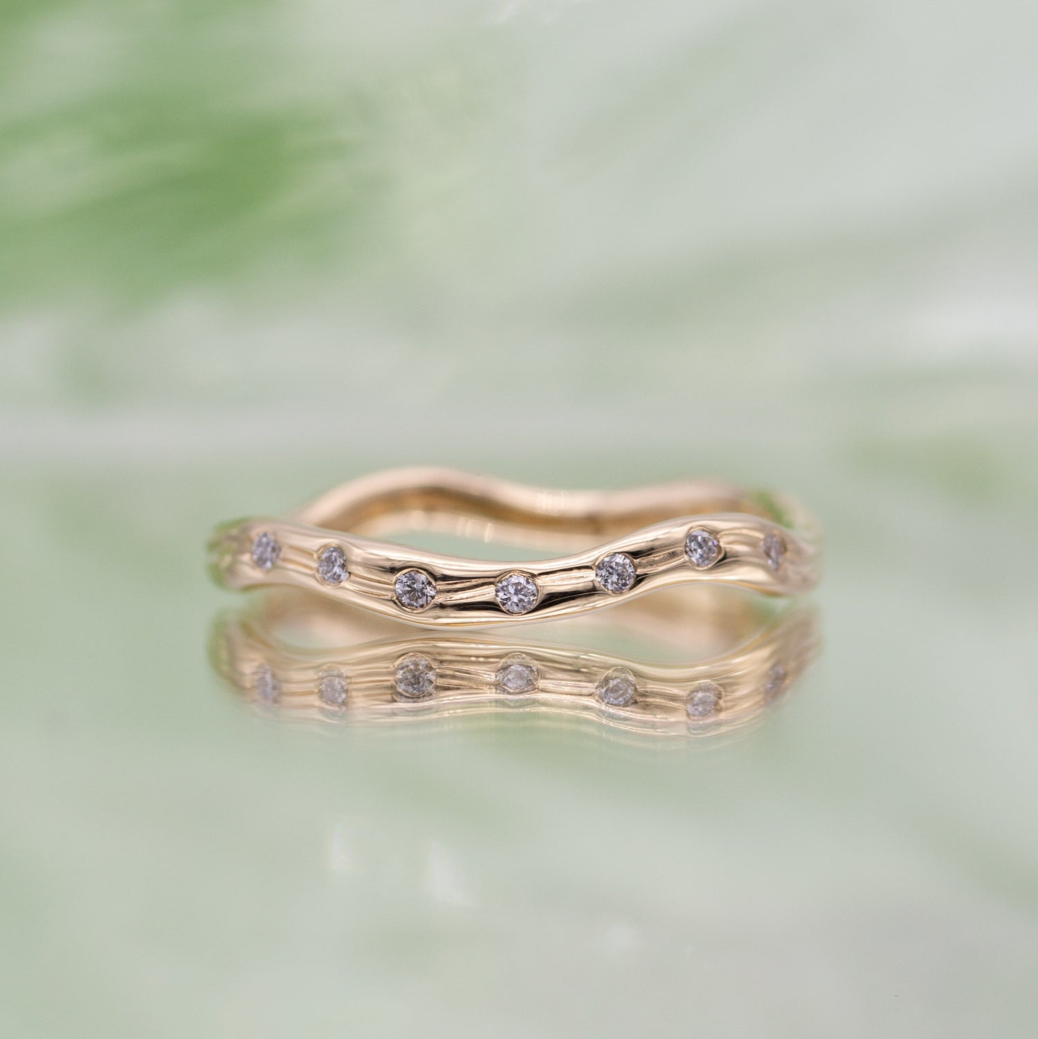 En ring i guld set forfra med syv diamanter på spejlende lysegrøn baggrund. Ringen er formgivet organisk og bølger elegant.