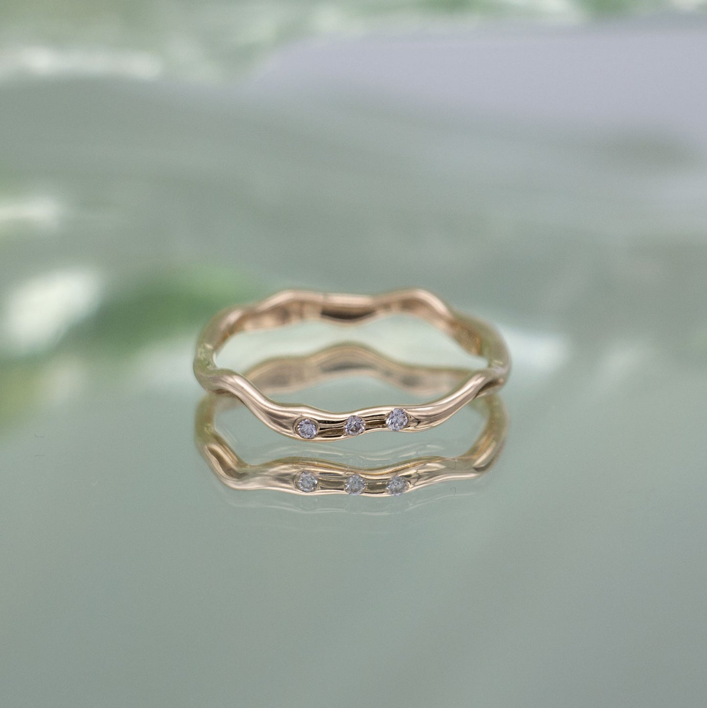 En ring i guld med tre diamanter ligger på en spejlende overflade med grønlige nuancer.  Ringen har tre fairtrade diamanter og er organisk formgivet.