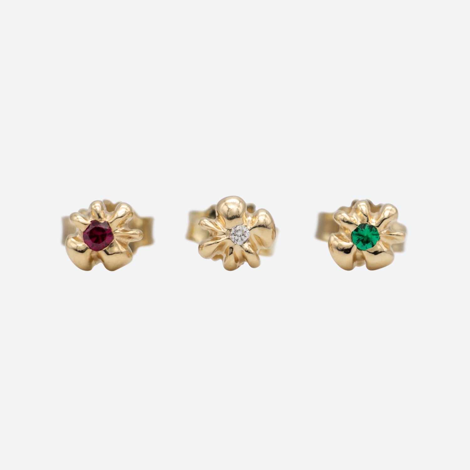 Tre ørestikker i guld formet som en blomst med henholdsvis rubin, diamant og smaragd set forfra på hvid baggrund. Design af Michell Liljefelt