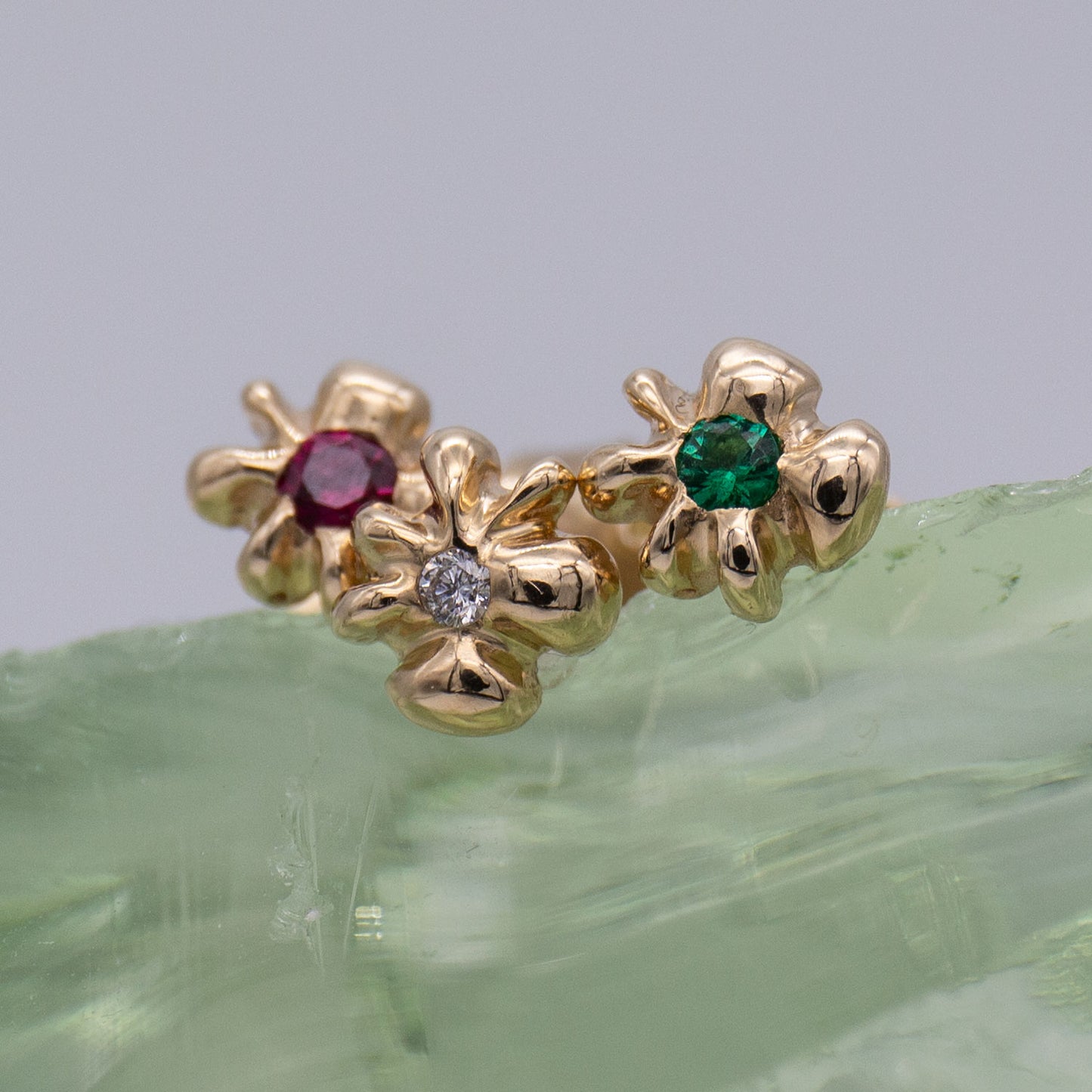 Detalje af tre ørestikker i guld formet som en blomst med henholdsvis en rubin, en diamant og en smaragd i midten siddende på en klump glas. Design af Michell Liljefelt