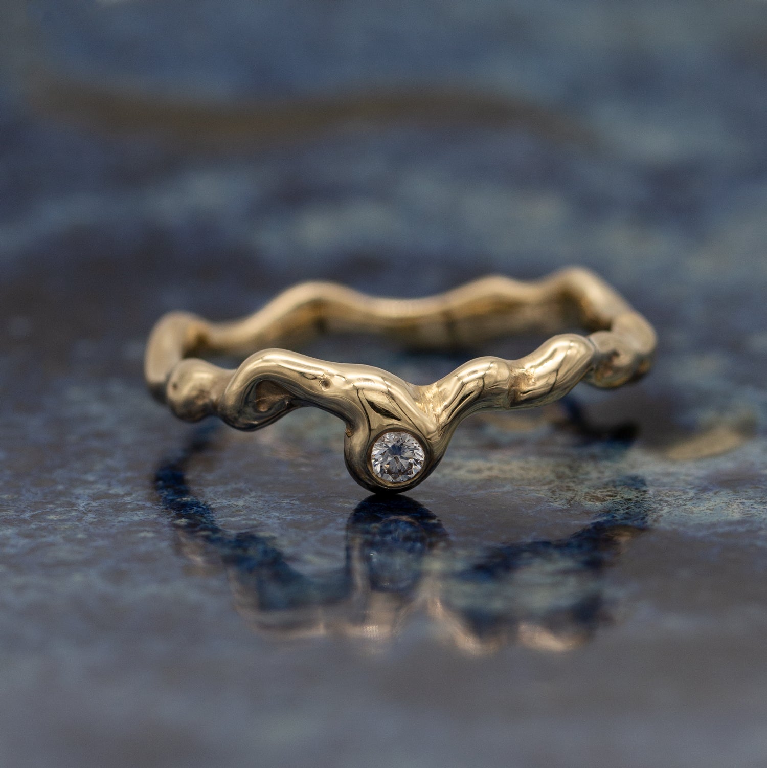 Organisk design ring i guld med en diamant på blå keramisk glasur baggrund der spejler blidt. 