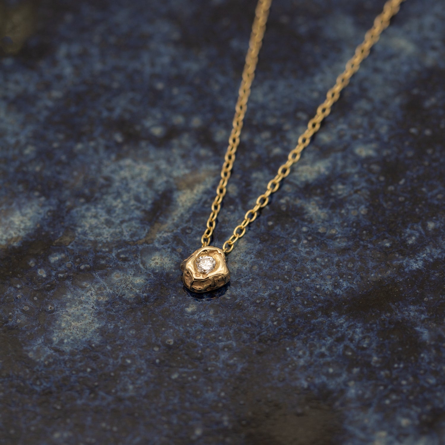 Halskæde i guld med vedhæng ligger på en keramisk overflade med mørke blå glasur. Halskæden i guld har et vedhæng der er formgivet organisk som et stykke fjeld, med en stor hvid diamant i midten der glimter.